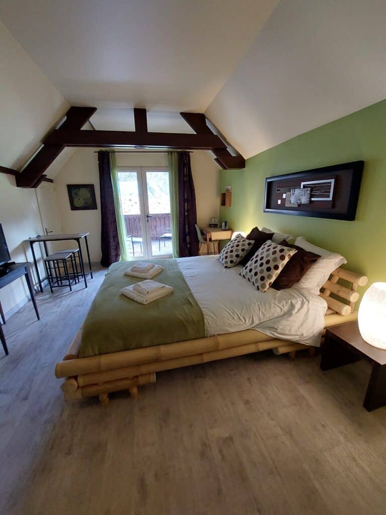 Chambre mansardée Zen avec lit en bambou, mur vert anis, poutres sombres. Couette blanche et serviettes. Coin-repas et balcon.
