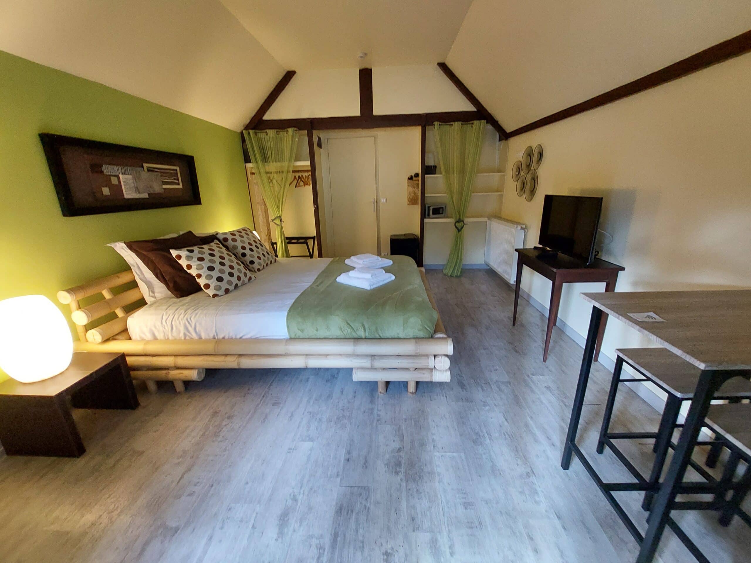 Chambre mansardée lumineuse, style zen. Lit en bambou, jeté vert olive et mur d'accent. Poutres sombres apparentes et coin repas moderne.
