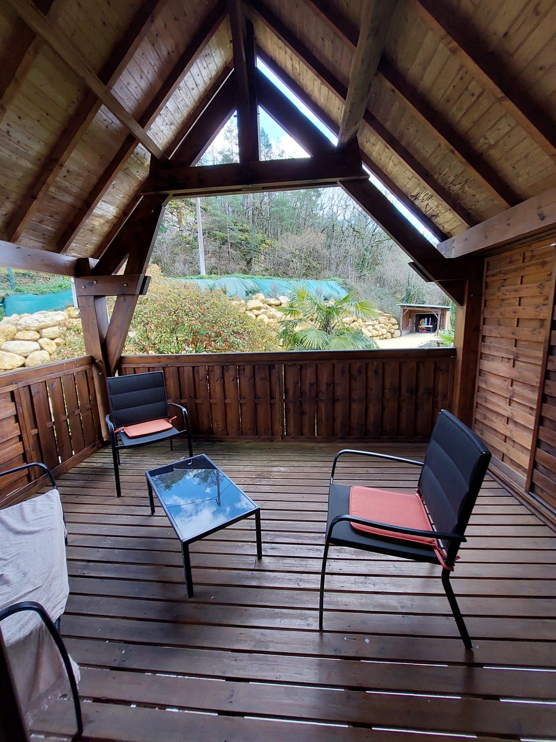Gazebo en bois rustique avec plancher à lattes, chaises noires et coussins corail. Vue ouverte sur une colline boisée et des rochers.
