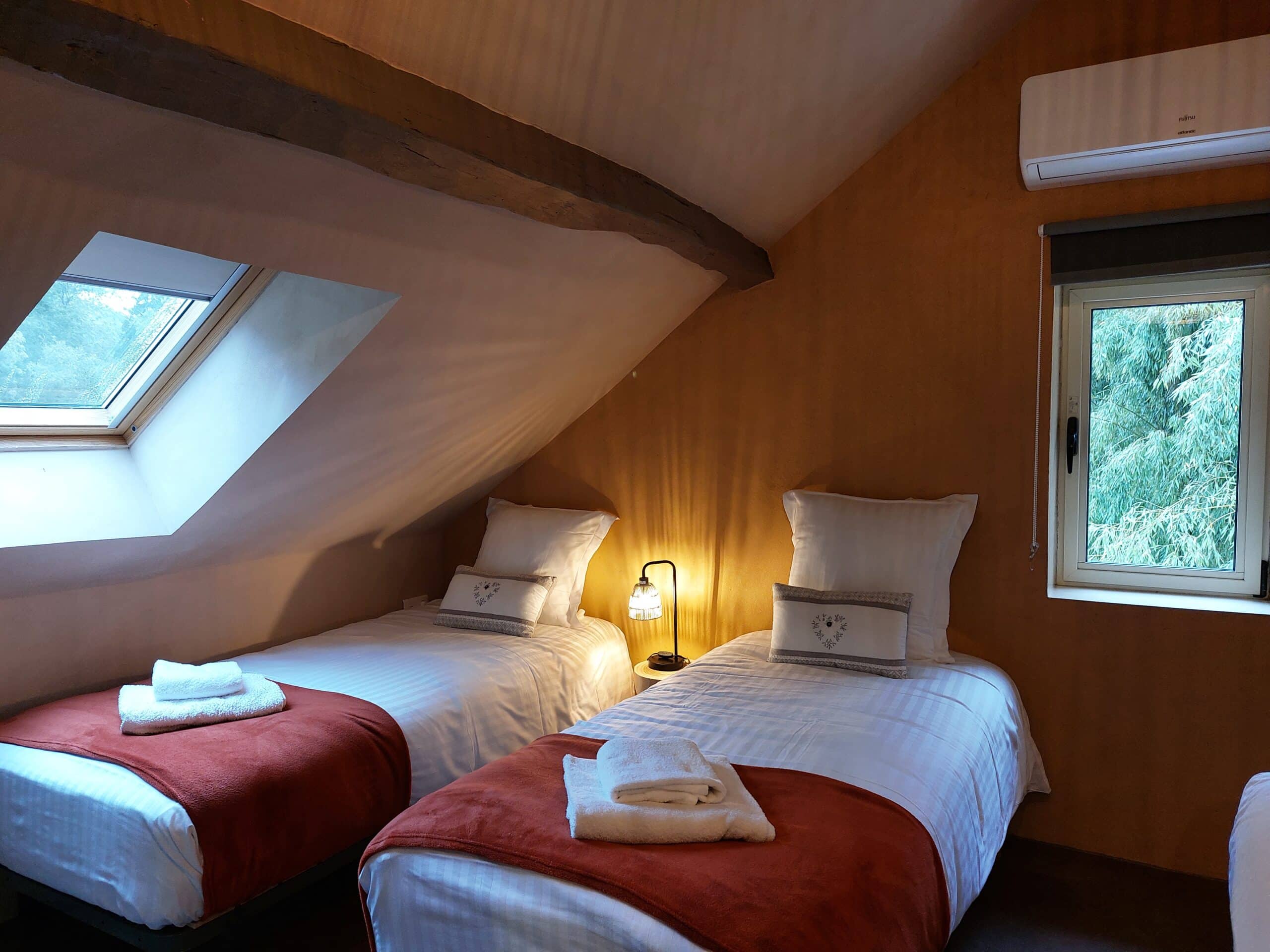 Chambre mansardée avec lits jumeaux, jetés rouille, lucarne et poutre. Climatisation Fujitsu Atlantic visible sur mur ocre.
