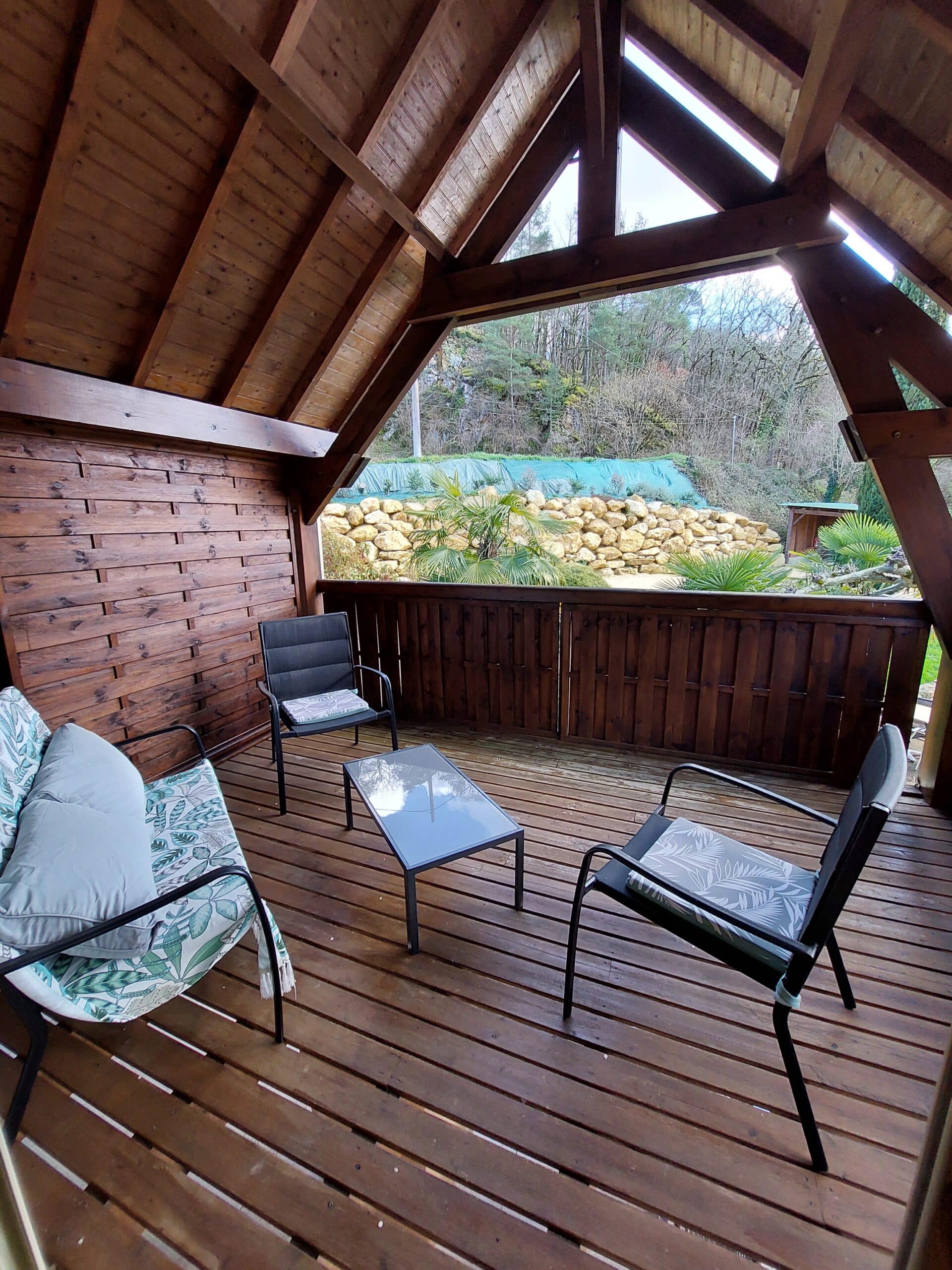 Terrasse couverte en bois avec salon de jardin (chaises, table). Structure rustique en poutres. Vue sur mur de pierres, palmiers et nature environnante.