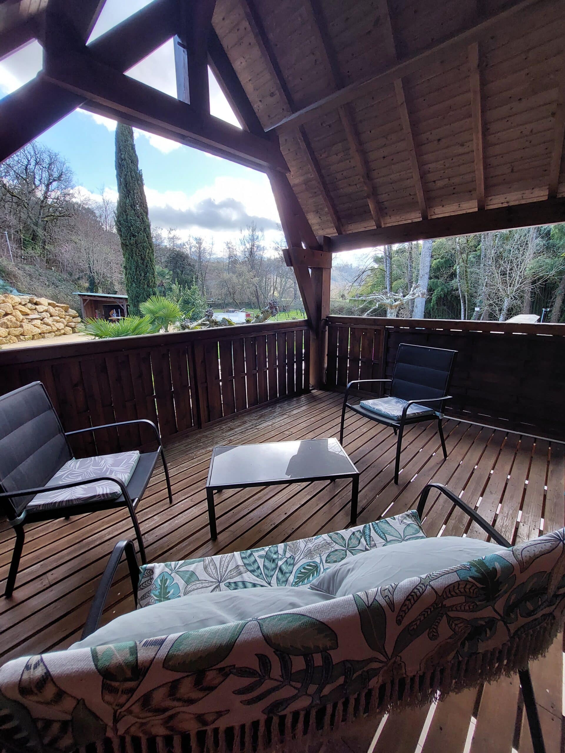 Terrasse en bois couverte aménagée en salon de jardin. La vue donne sur une rivière, une colline boisée et un grand cyprès.