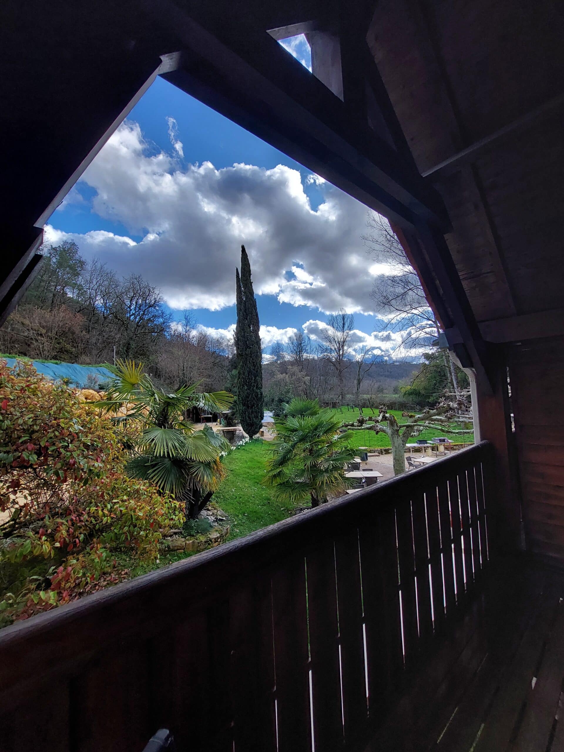 Balcon en bois sombre. Cadre triangulaire sur jardin verdoyant avec palmiers et cyprès élancé. Ciel bleu intense et nuages dramatiques.