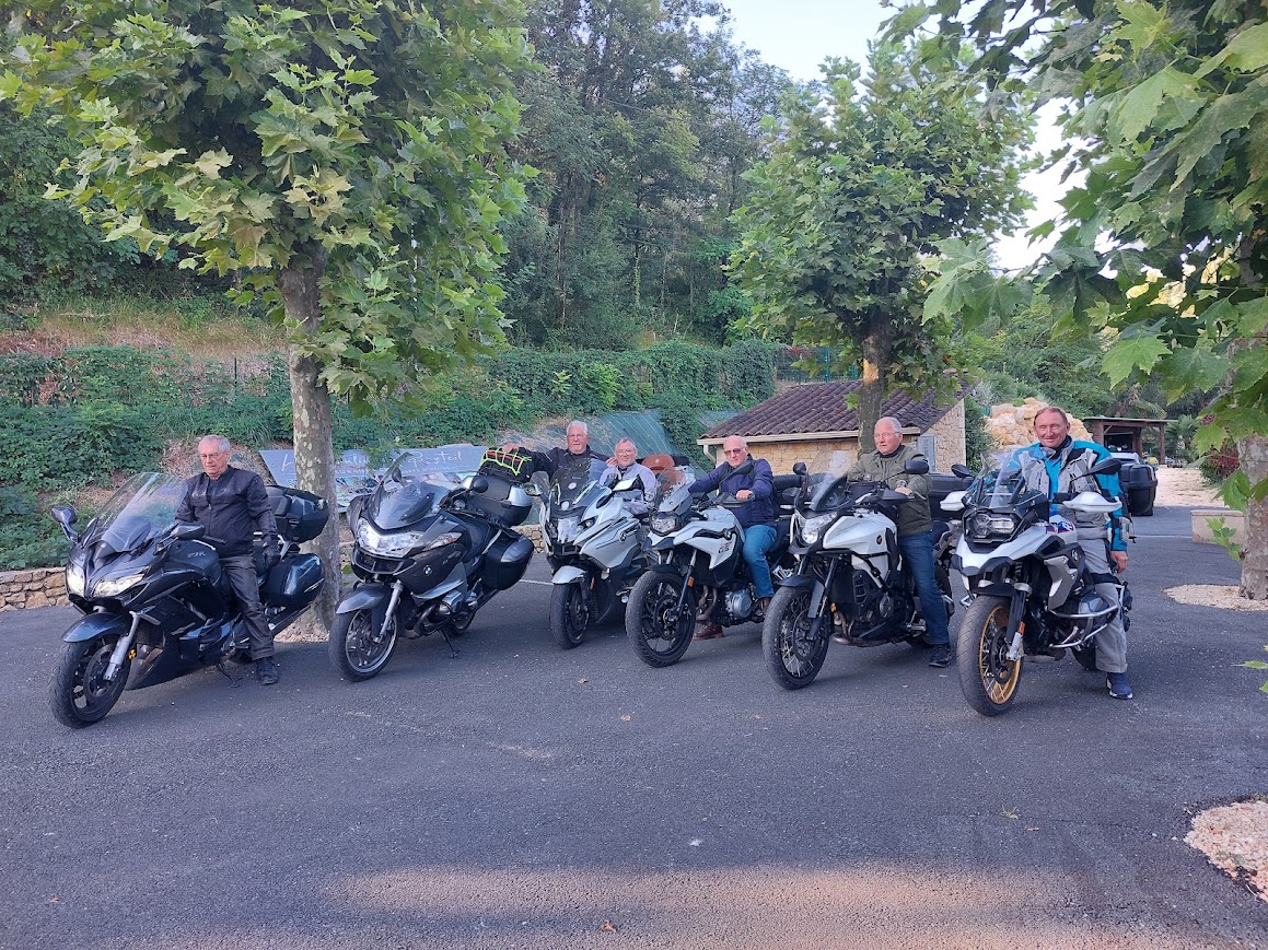 Six motards d'âge mûr posent avec leurs motos de tourisme et aventure, majoritairement BMW, dans un cadre verdoyant.