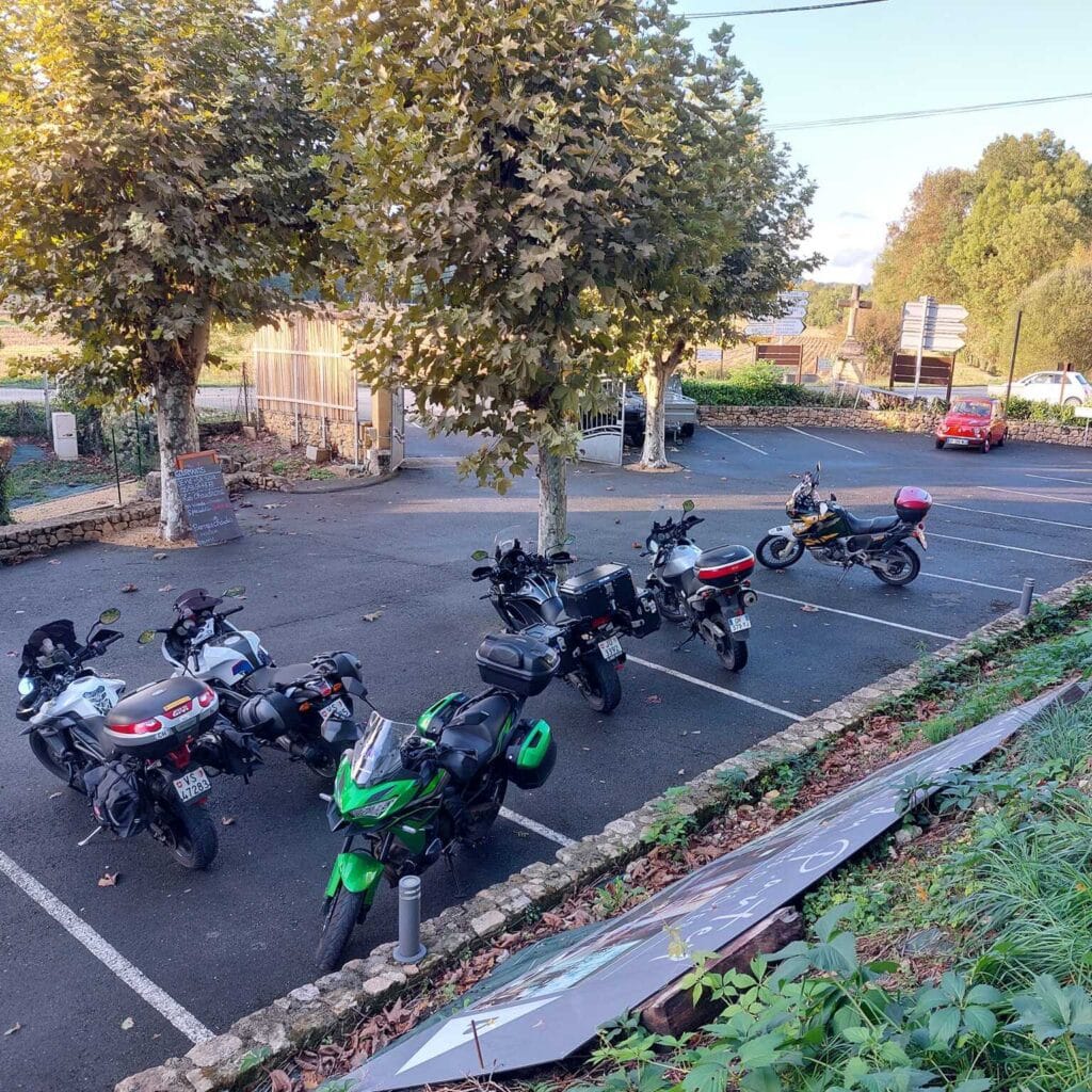 Motos de voyage garées : Pause étape sous les platanes Sept motos de tourisme (vert, blanc, noir) avec bagages garées sous des platanes. Lumière dorée, parking près d'un menu sur ardoise.