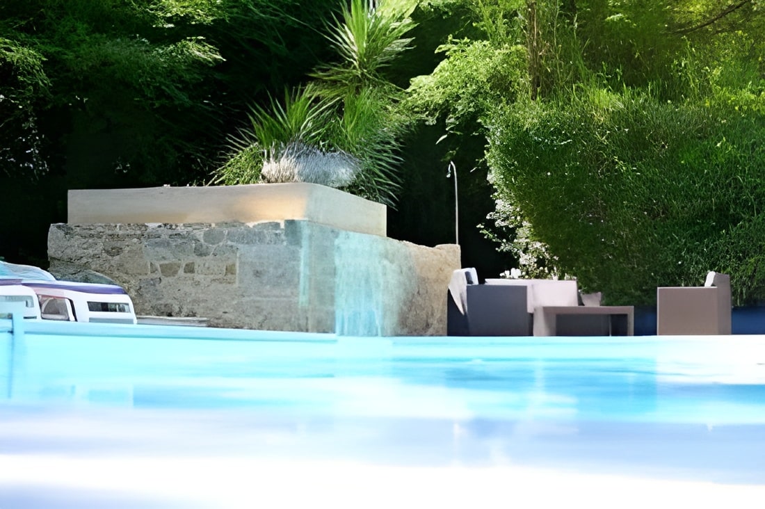 Piscine contemporaine reflétant le ciel, bordée par un salon extérieur moderne et un mur de pierre rustique. Végétation verte et luxuriante.